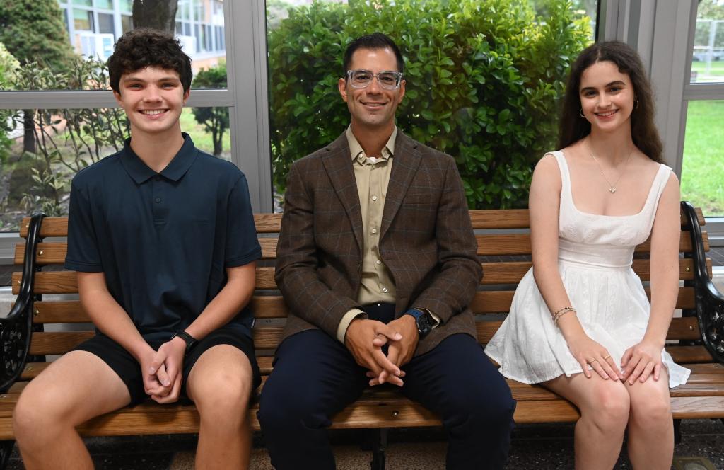 Three people sit together on a wooden park bench, smiling for the camera indoors.
